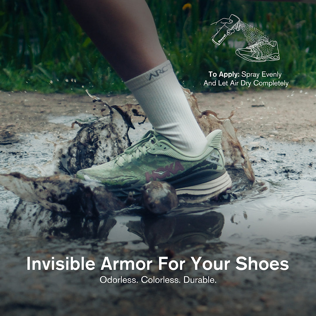 A green HOKA running sneaker with white crew socks stepping through a muddy puddle with water and dirt splashing around it. An illustrated icon in the top right shows the Jason Markk Repel spray being applied to a shoe with the instruction: "To Apply: Spray Evenly And Let Air Dry Completely." Text overlay reads: "Invisible Armor For Your Shoes – Odorless. Colorless. Durable."