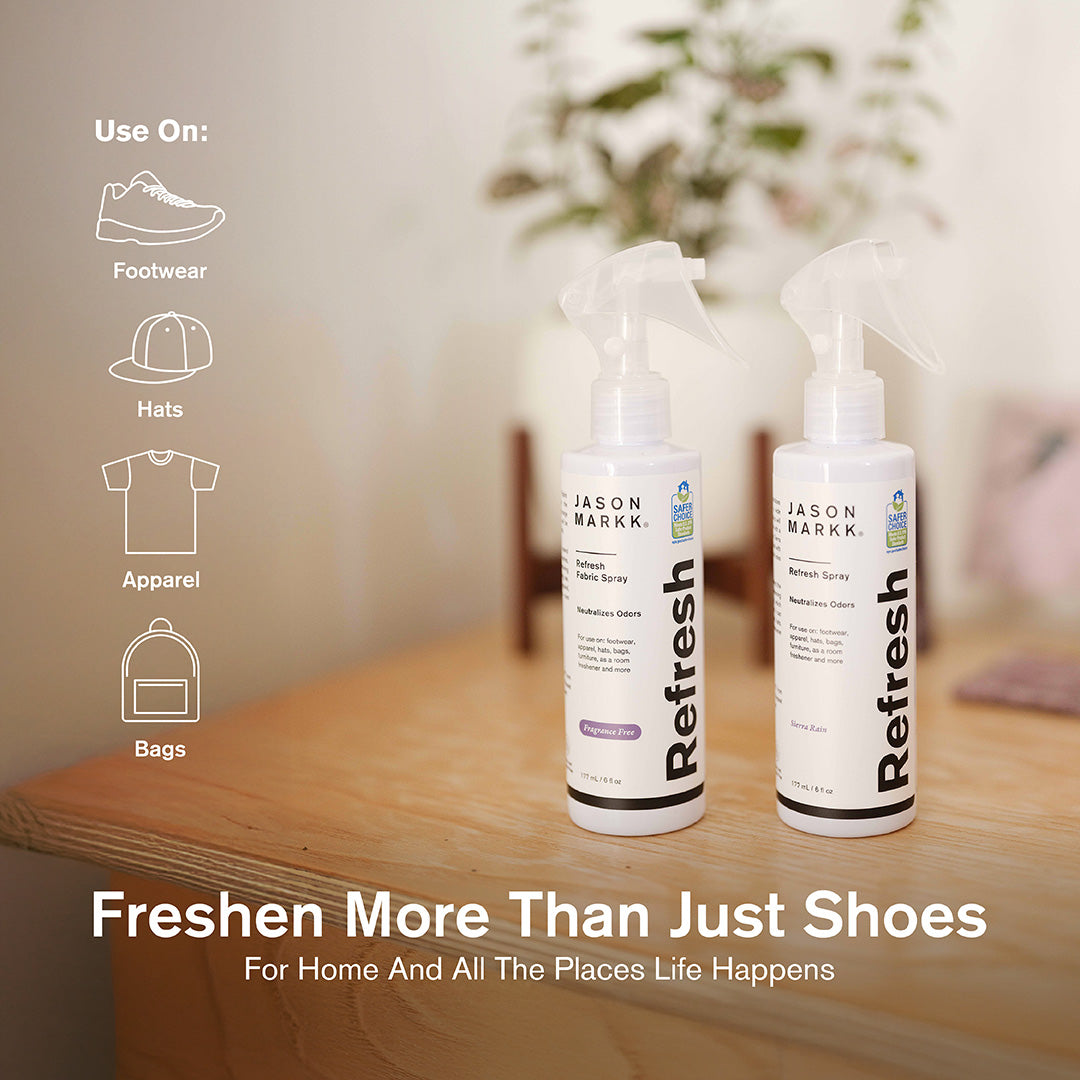 Two 6 fl oz Jason Markk Refresh odor neutralizing spray bottles displayed side by side on a wooden nightstand in a softly lit home setting. The left bottle is labeled Refresh Fabric Spray in Fragrance Free and the right is Refresh Spray in Sierra Rain scent, both EPA Safer Choice certified. Icons on the left indicate compatible uses: Footwear, Hats, Apparel, and Bags. Text overlay reads: "Freshen More Than Just Shoes – For Home And All The Places Life Happens."