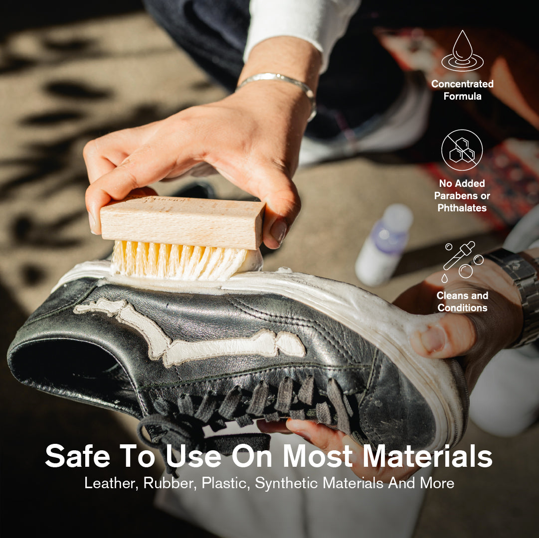 A person wearing a silver bracelet scrubbing a heavily soiled black leather sneaker with a Jason Markk Durables wooden stiff bristle brush, with a bottle of Jason Markk Deep Cleaning Solution visible in the background. Three feature icons on the right highlight: Concentrated Formula, No Added Parabens or Phthalates, and Cleans and Conditions. Text overlay reads: "Safe To Use On Most Materials – Leather, Rubber, Plastic, Synthetic Materials And More."